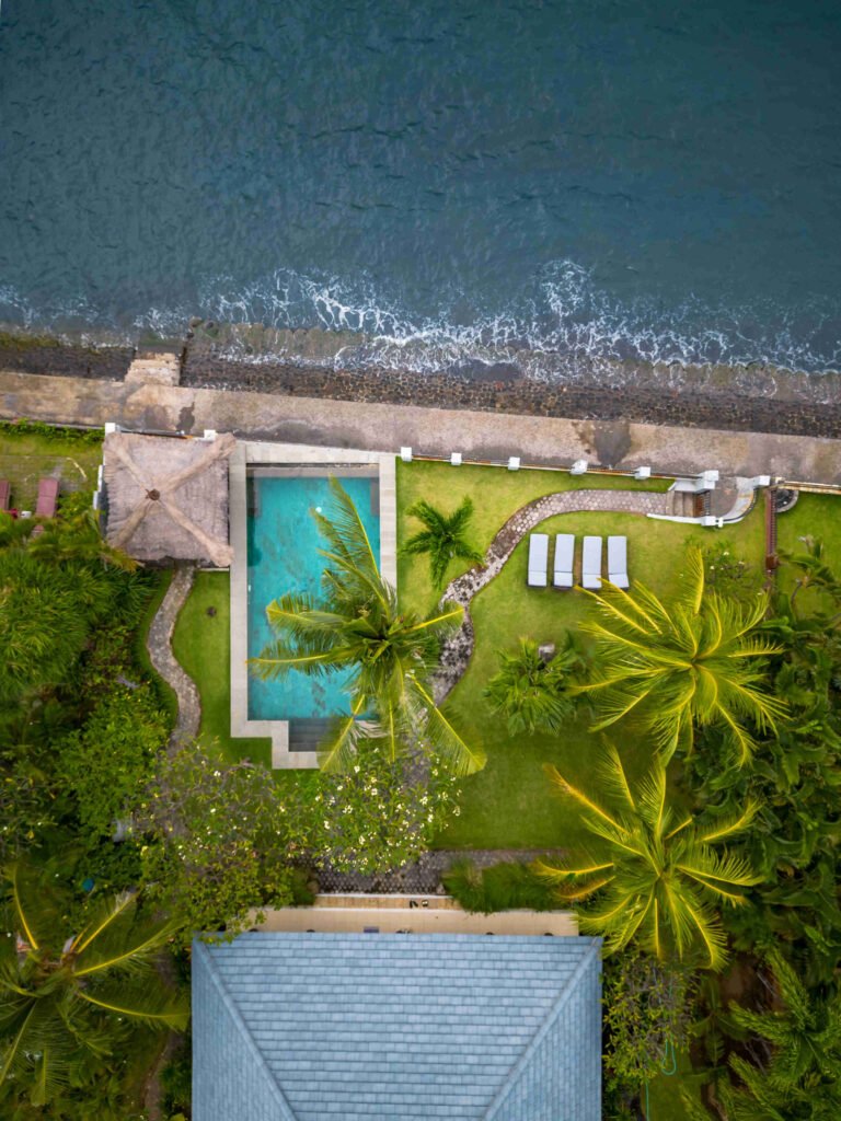 Overhead drone view of Villa Seaview Bukti, with waves breaking on the black rock seawall beside the turquoise pool, lawn and thatched bale
