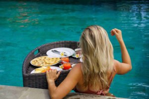 A guest in the pool at Villa Seaview Bukti beside a floating breakfast tray of pancakes, sliced fruit and fresh juice in the turquoise water