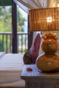 Close-up of a lit bamboo bedside lamp with a shell-trimmed base at Samastiti by the Beach in Lovina, canopy net and green balcony blurred behind