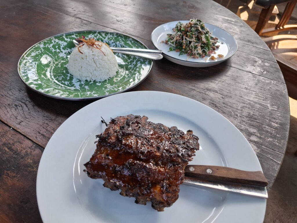 Grilled pork ribs with steamed rice and urab greens on a teak table at Dapur Bali Mula, Les village